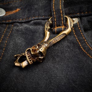 May include: A bronze-colored metal keychain with a skull design. The skull is detailed with visible teeth and eye sockets. The keychain is attached to a dark denim fabric with orange stitching.