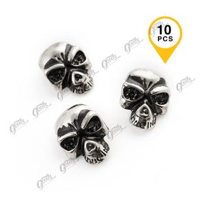Può includere: Tre perline a forma di teschio color argento con dettagli del viso sono mostrate su sfondo bianco. Le perline hanno orbite scure e una superficie strutturata. Un'etichetta gialla indica "10 PCS".