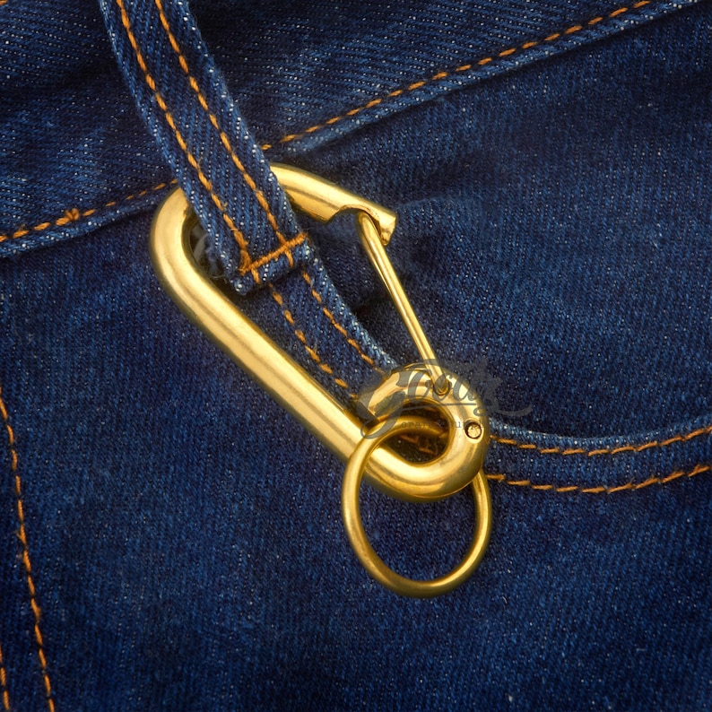 Puede incluir: Primer plano de un mosquet&oacute;n dorado con un anillo circular, sujeto a una tela vaquera azul con costuras naranjas.