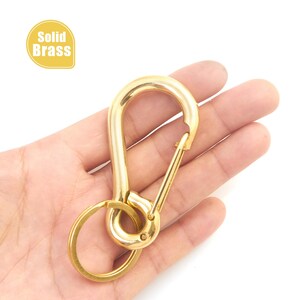 以下が含まれることがあります： 金色の真鍮製ソリッドキーチェーンカラビナ。スプリング式のゲートと小さなキーリングが付いています。「Solid Brass」の文字が黄色の楕円形で表示されています。