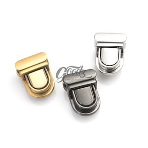 May include: Three metal purse clasps in gold, silver, and gunmetal finishes. Each clasp has a rectangular top and a U-shaped latch. The clasps are displayed on a white background, showcasing their different colors and designs.