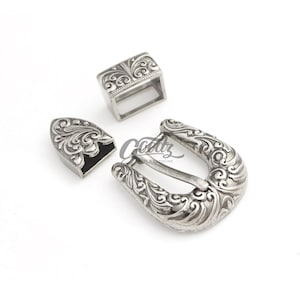 May include: A set of three silver-colored metal belt hardware pieces. The set includes a rectangular loop, a decorative tip, and a horseshoe-shaped buckle, all with an ornate, swirling floral design.