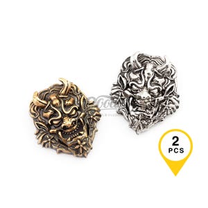 May include: Two metal Oni mask charms. One is bronze-colored, the other is silver-colored. The charms are detailed with horns, fangs, and intricate facial features. A yellow graphic indicates that there are two pieces.