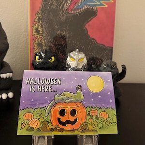 May include: A Halloween-themed greeting card with the text "HALLOWEEN IS HERE..." features a cartoon alligator resting on a jack-o'-lantern. The card is propped up in front of Godzilla figurines and a Godzilla poster.
