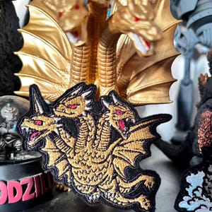 May include: An embroidered patch featuring a three-headed golden dragon with red eyes and sharp teeth, set against a black background. The design showcases detailed scales, wings, and horns, capturing the creature in a dynamic, multi-headed pose.