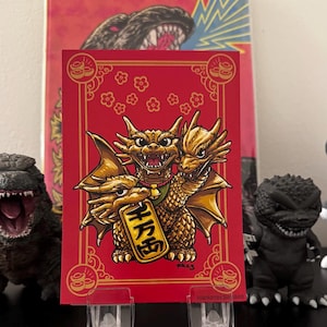 May include: A red and gold card with a three-headed golden dragon holding a gold charm with Chinese characters. The dragon has three heads, each with a different expression. The card is decorated with gold flowers and a gold border.