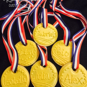 May include: Six gold-colored cookies decorated with the names "Gabriel", "Aiden", "Gio", "Isabella", "Julia", and "Alex". Each cookie has a red, white, and blue ribbon attached.