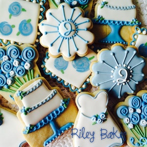 May include: A collection of blue and white decorated sugar cookies featuring wedding-themed designs, including a cake, a bouquet of flowers, and a dress. The cookies are decorated with blue and white icing and have a delicate, hand-painted look. The text "Riley Bakes" is visible in the bottom right corner of the image.