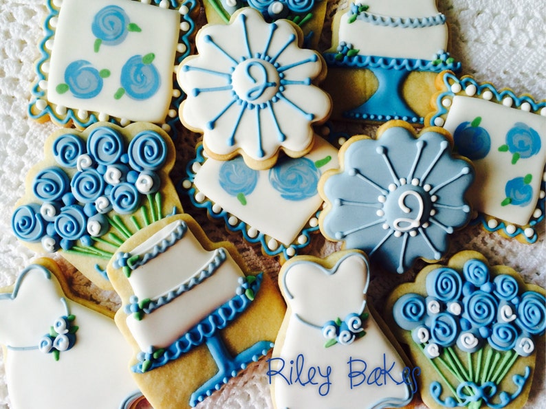 May include: A collection of blue and white decorated sugar cookies featuring wedding-themed designs, including a cake, a bouquet of flowers, and a dress. The cookies are decorated with blue and white icing and have a delicate, hand-painted look. The text "Riley Bakes" is visible in the bottom right corner of the image.