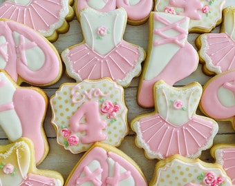Op de afbeelding: Een set roze en witte gedecoreerde suikerkoekjes met ballerina tutu's, balletten en het nummer vier. De koekjes zijn versierd met gouden stippen en roze bloemen. Een koekje heeft de naam "Eva" erop.