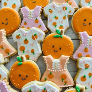 May include: A collection of decorated sugar cookies featuring orange slices, baby onesies, and dresses. The cookies are decorated with orange and pink icing and have a whimsical, baby shower theme. The text "Riley Bakes Custom Sweets & More" is visible in the bottom right corner of the image.