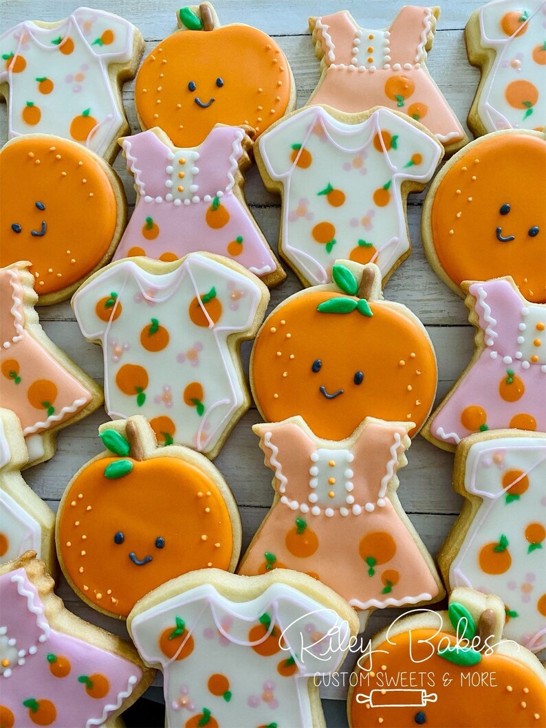 May include: A collection of decorated sugar cookies featuring orange slices, baby onesies, and dresses. The cookies are decorated with orange and pink icing and have a whimsical, baby shower theme. The text "Riley Bakes Custom Sweets & More" is visible in the bottom right corner of the image.