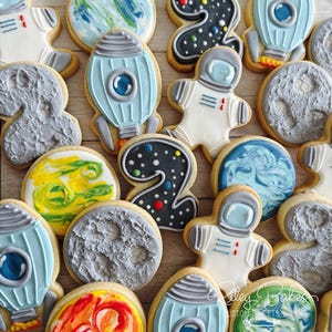 May include: A collection of space-themed sugar cookies featuring iced designs of rockets, astronauts, planets with marbled icing, and the number two decorated with colorful dots on a dark background, arranged in a scattered pattern.