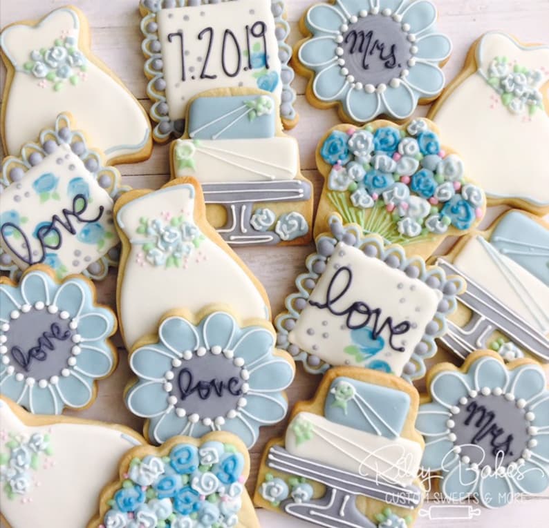May include: Assortment of decorated sugar cookies with blue and white icing. The cookies feature various designs including wedding dresses, flowers, cakes, and the words "love" and "Mrs.".