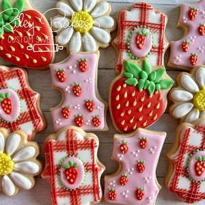 May include: Assortment of decorated sugar cookies. Designs include strawberries, daisies, and plaid patterns. Some cookies are shaped like the number one, decorated with small strawberries. The cookies are iced in red, pink, white, and green.