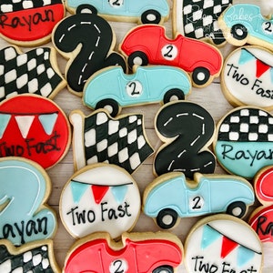 May include: A collection of decorated sugar cookies featuring a racing car theme. The cookies are decorated with checkered flags, racing numbers, and the words "Two Fast" and "Rayan".