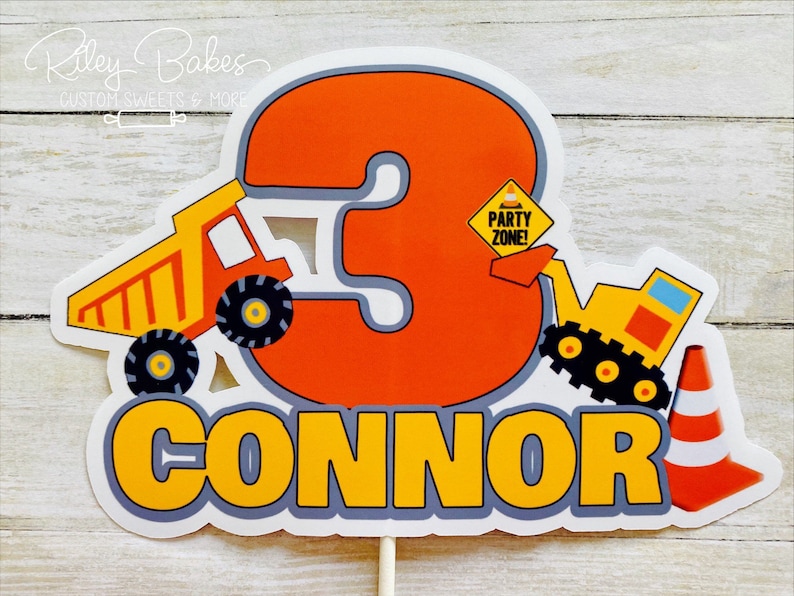 May include: Orange and yellow construction themed cake topper with the number 3, a dump truck, an excavator, and a traffic cone. The name Connor is written in yellow letters with a gray outline.