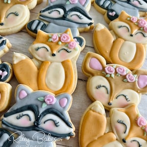 May include: Assortment of decorated sugar cookies featuring woodland animal designs. The cookies are decorated with royal icing and feature foxes, raccoons, and deer. The cookies are decorated with pink and white flowers.