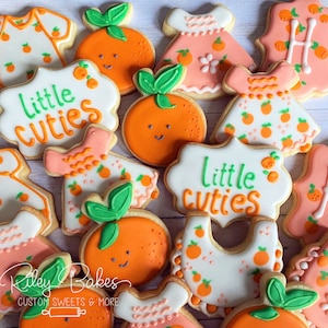 May include: Assortment of decorated sugar cookies with orange and pink frosting. The cookies feature various designs, including dresses, bibs, and the phrase "Little Cuties".