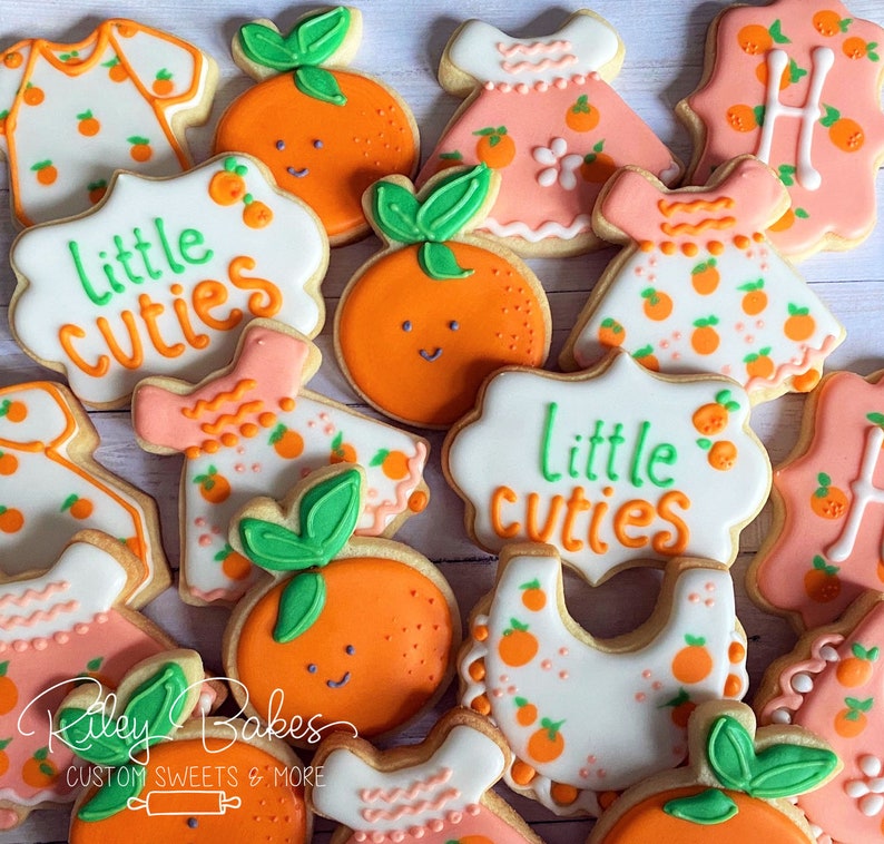 May include: Assortment of decorated sugar cookies with orange and pink frosting. The cookies feature various designs, including dresses, bibs, and the phrase "Little Cuties".