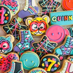 May include: A collection of decorated sugar cookies in various shapes, including stars, roller skates, and smiley faces. The cookies feature bright colors like pink, teal, and yellow, with black accents and the name "Quinn" on one cookie. The cookies are from Riley Bakes.