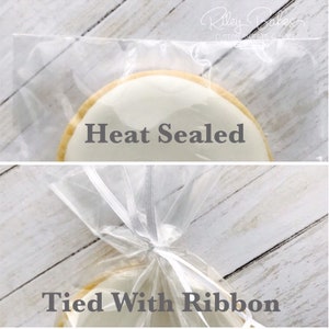 May include: A round cookie with white icing is heat sealed in a clear plastic bag and tied with a white ribbon. The text "Heat Sealed" and "Tied With Ribbon" are visible on the image. The cookie has a golden-brown edge.
