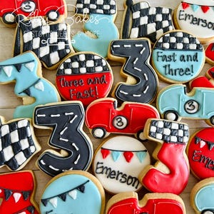 May include: A collection of decorated sugar cookies featuring a race car theme. The cookies are decorated with red, blue, black, and white icing. Some cookies feature the number three, while others have checkered flags, race cars, and the name "Emerson".