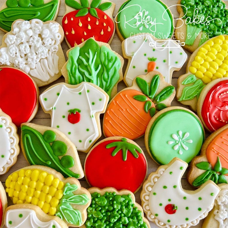May include: A collection of decorated sugar cookies featuring various vegetables and fruits, including a strawberry, a carrot, a tomato, corn, peas, lettuce, and broccoli.  The cookies are decorated with white icing and green, red, yellow, and orange food coloring.  The cookies are arranged in a random pattern on a wooden surface.  The text "Riley Bakes Custom Sweets & More" is visible in the top right corner of the image.