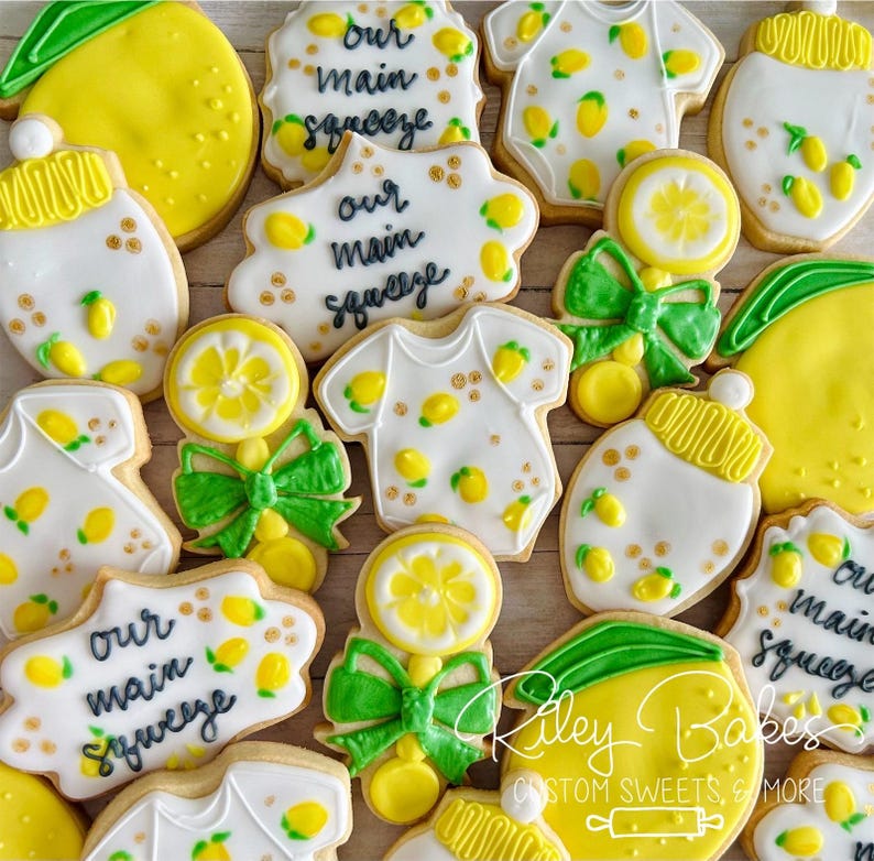Pu&ograve; includere: Una collezione di biscotti di zucchero decorati con un tema di limone. I biscotti sono decorati con glassa gialla e verde e presentano vari disegni, tra cui limoni, body per neonati e sonagli. Il testo "Our Main Squeeze" &egrave; scritto su diversi biscotti.