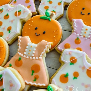 May include: A collection of decorated sugar cookies featuring orange and floral designs. The cookies are shaped like onesies, dresses, and oranges. The cookies are decorated with white, pink, and orange icing. The text "Riley Bakes Custom Sweets & More" is visible in the bottom right corner of the image.