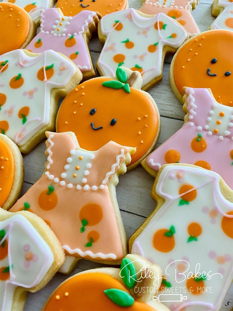 May include: A collection of decorated sugar cookies featuring orange and floral designs. The cookies are shaped like onesies, dresses, and oranges. The cookies are decorated with white, pink, and orange icing. The text "Riley Bakes Custom Sweets & More" is visible in the bottom right corner of the image.