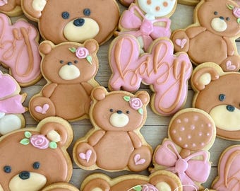 May include: An assortment of decorated sugar cookies featuring teddy bears, a rattle, and the word "baby" in pink icing. The cookies are decorated with pink and brown icing and have a rustic wood background.