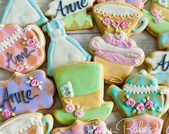 May include: A collection of Alice in Wonderland themed sugar cookies decorated with various characters and designs. The cookies feature a teapot, a teacup, a Mad Hatter's hat, and a dress. The cookies are decorated with pastel colors and intricate details. The cookies are labeled with the name "Anne".