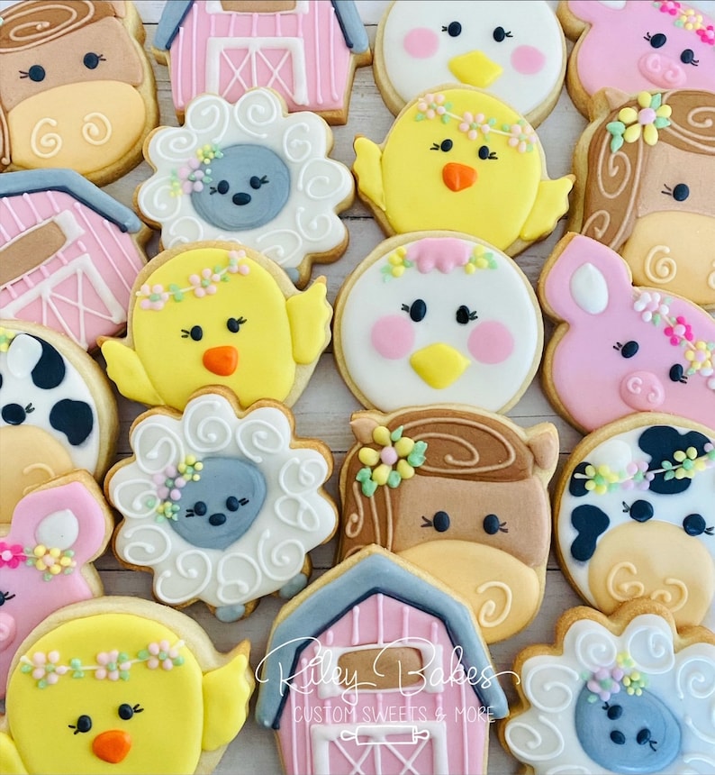 May include: A collection of farm animal themed sugar cookies decorated with colorful frosting. The cookies feature a variety of animals, including cows, pigs, chickens, horses, and sheep. The cookies are decorated with flowers, swirls, and other details. The cookies are arranged on a white surface. The text "Riley Bakes Custom Sweets & More" is visible in the bottom right corner of the image.