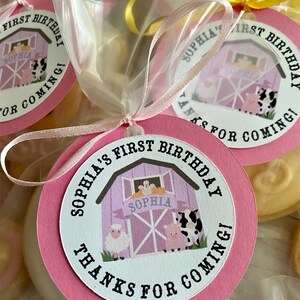 May include: Pink and white favor bags with a round pink tag that says "Sophia's First Birthday Thanks for Coming!" with a cartoon barn and farm animals. The tag is tied with a pink ribbon.