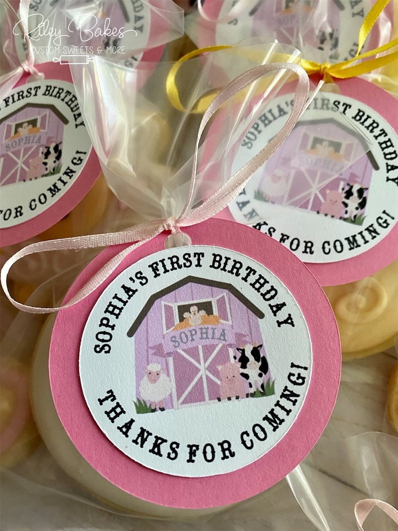 May include: Pink and white favor bags with a round pink tag that says "Sophia's First Birthday Thanks for Coming!" with a cartoon barn and farm animals. The tag is tied with a pink ribbon.