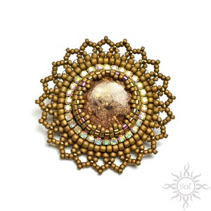 Peut inclure: Broche circulaire faite à la main, ornée d'une pierre centrale marron translucide avec des inclusions de feuilles d'or. La broche est entourée de petites perles dorées et de cristaux irisés, avec un bord extérieur perlé décoratif.