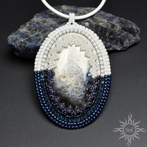 Puede incluir: Collar con colgante ovalado y cordón blanco. El colgante presenta una piedra central con patrones grises y blancos, enmarcada por cuentas blancas y azul marino. El diseño es intrincado, con una mezcla de texturas y colores, creando una joya única.