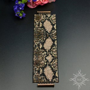 Könnte beinhalten: Ein schwarzes und goldenes Armband mit Schlangenhautmuster. Das Armband hat zwei Metallverschlüsse.