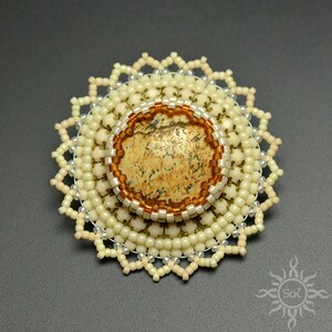Peut inclure: Broche circulaire faite à la main, avec une pierre centrale tachetée. La pierre est encadrée de perles marron et blanches, entourée de couches de perles crème. Le bord extérieur présente un motif en étoile délicat, pour une pièce d'art portable unique.