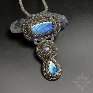 Puede incluir: Un collar de cuentas plateadas con un gran colgante de labradorita azul. El colgante está rodeado de cuentas plateadas más pequeñas y piedras azules. El collar está sobre un fondo gris oscuro.