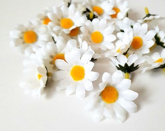 May include: A bunch of white daisy flowers with yellow centers. The flowers are arranged in a pile on a white surface.