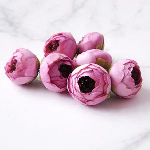 Puede incluir: Un grupo de ocho capullos de flores de peonía rosas artificiales con centros oscuros. Las flores artificiales están dispuestas sobre una superficie de mármol blanco, mostrando sus pétalos en capas y su apariencia realista. Ideal para manualidades o decoración.