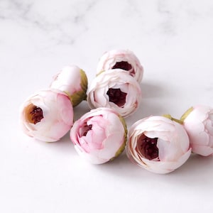 May include: A collection of delicate, artificial peony flower heads. The petals are a soft pink, with a darker, burgundy center. The flowers are arranged on a white marble surface, perfect for crafting or decorative purposes.