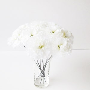 May include: A bouquet of white silk flowers arranged in a clear glass vase. The flowers are on thin wire stems.