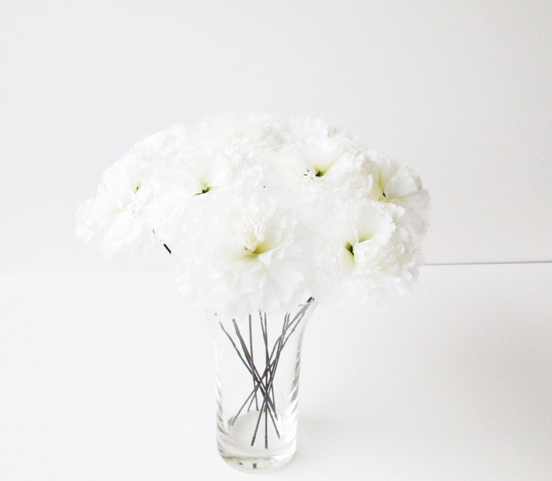 May include: A bouquet of white silk flowers arranged in a clear glass vase. The flowers are on thin wire stems.