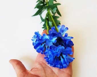 May include: A bouquet of blue artificial flowers with green stems. The flowers are arranged in a hand.