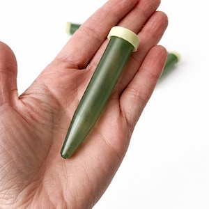 Puede incluir: Una mano sostiene un tubo de agua floral de plástico verde con una tapa amarillo claro. El tubo es cónico y tiene una pequeña abertura en la parte inferior. Dos tubos adicionales son visibles en el fondo, sobre un fondo blanco.