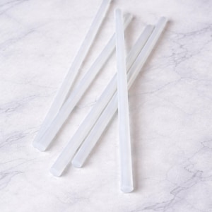 May include: Several translucent, white glue sticks are scattered on a white and grey marble surface. These cylindrical glue sticks, with flat ends, are designed for use in a glue gun, suitable for crafts and DIY tasks.