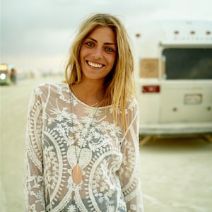 May include: A woman wearing a white lace top with a long pendant necklace. The top is sheer and has a floral pattern. The woman is smiling and looking at the camera.
